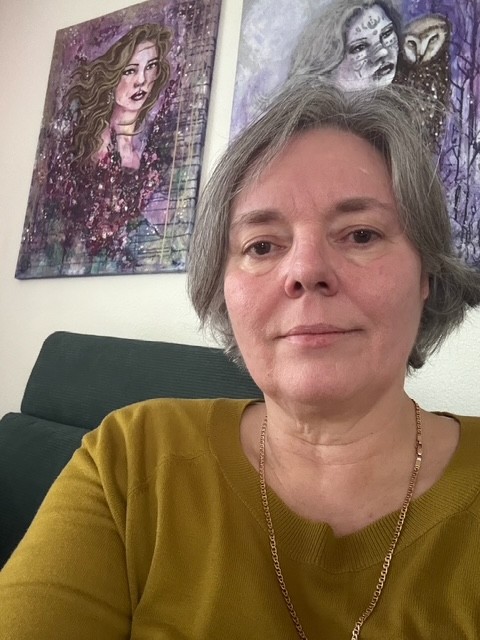 Carolyn Pini am grünen Sofa, zwei ihrer Gemälde im Hintergrund